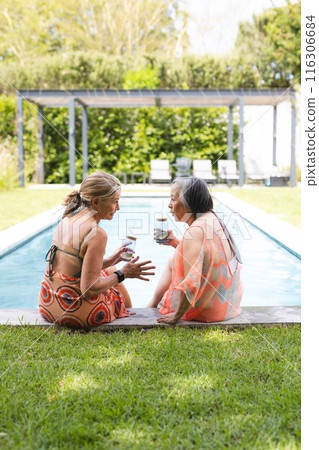 Outdoors, diverse senior female friends sitting by pool, chatting, holding drinks, copy space 116306684