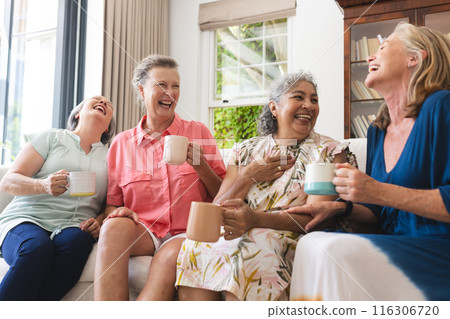 Diverse senior female friends holding mugs, laughing together at home Diverse senior female friends holding mugs, laughing together at home 116306720