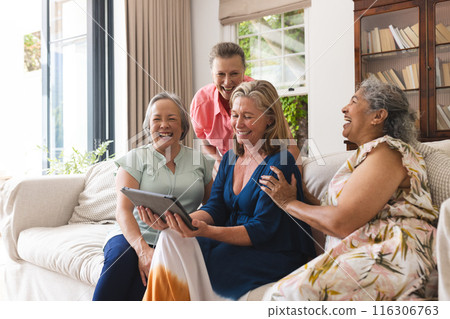 Diverse senior female friends laughing at home, one holding tablet Diverse senior female friends laughing at home, one holding tablet 116306763