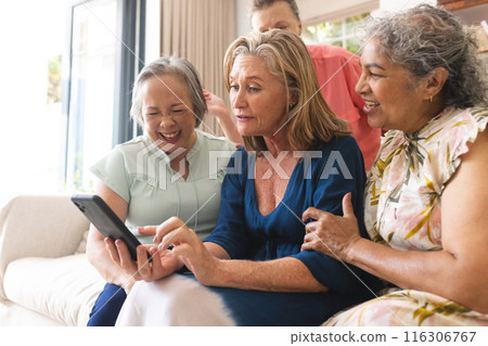 Diverse senior female friends laughing at home, looking at tablet 116306767