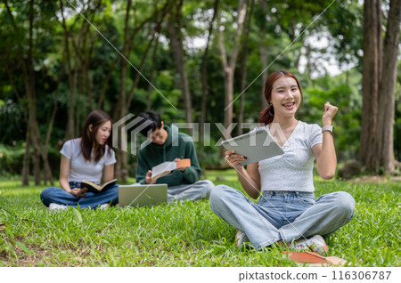 A positive Asian female college student sits on the grass in the park with her digital tablet. A positive Asian female college student sits on the grass in the park with her digital tablet. 116306787