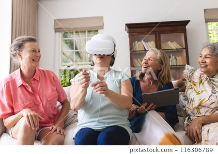 Diverse senior female friends trying on VR headset at home, laughing together 116306789