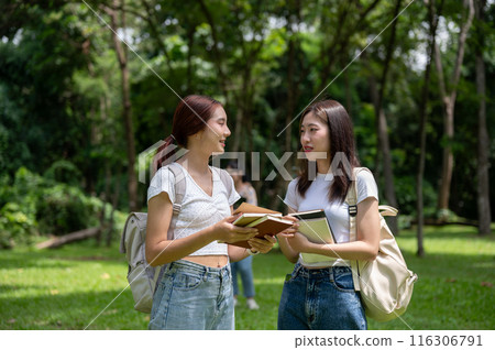 Two happy Asian female college students with backpacks are talking while walking together in park. 116306791