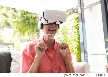 A Caucasian senior woman wearing virtual reality headset, laughing at home 116306811
