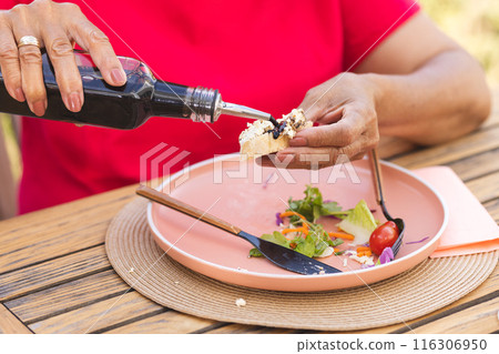 Senior biracial woman pouring dressing on salad outdoors, wearing red 116306950