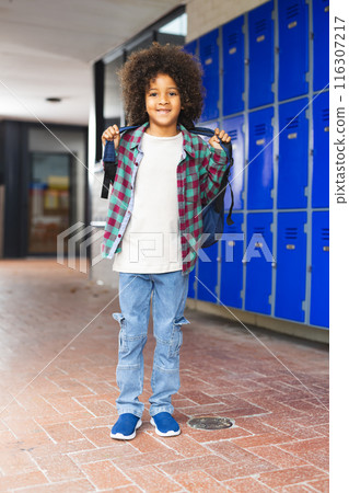 In school, young biracial boy wearing a backpack standing by lockers 116307217