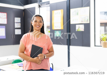 In school, young biracial female teacher holding a tablet, smiling, with copy space, in classroom 116307219