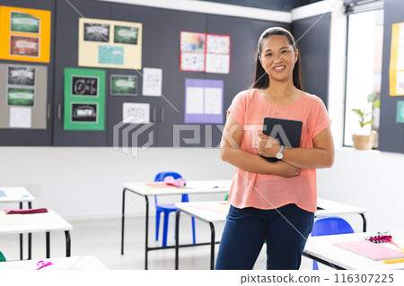 In school classroom, young biracial female teacher holding a tablet stands smiling with copy space 116307225