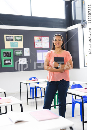 In school, young biracial female teacher holding a tablet stands smiling in the classroom 116307231