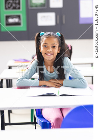 In school, young biracial girl with long, dark hair is sitting at a desk in a classroom 116307249