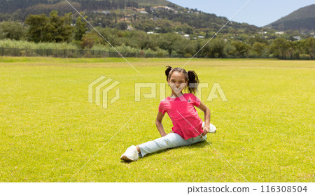 Biracial girl with pigtails is sitting on the grass in a park with copy space 116308504