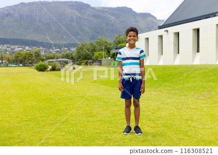 Biracial boy stands smiling on a sunny grass field, mountains in the background, with copy space 116308521
