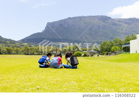 Three students are sitting on the grass, studying with a mountain in the background 116308522