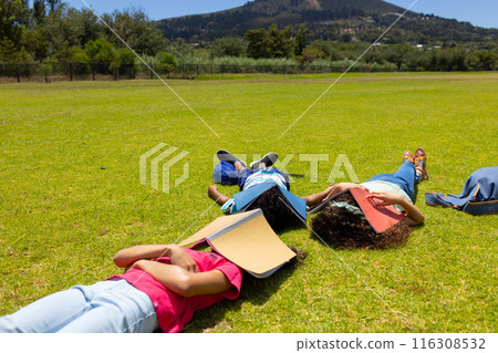 Three children are lying on the grass with books beside them, facing away from the camera 116308532