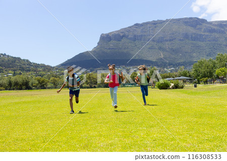 Three biracial children run across grassy field, mountains behind 116308533