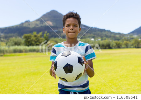 Biracial boy holds a soccer ball, standing in a sunny field with mountains behind 116308541