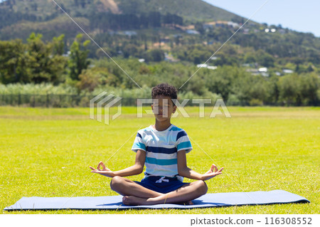 Biracial boy meditates on a yoga mat in a sunny park with copy space 116308552