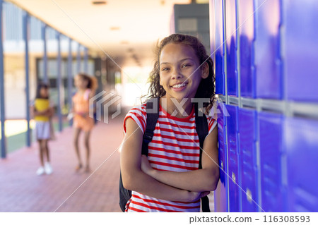 Biracial girl with curly brown hair smiles, leaning against blue lockers in school 116308593