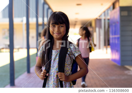 Biracial girl with long dark hair smiles in school, holding her backpack straps 116308600