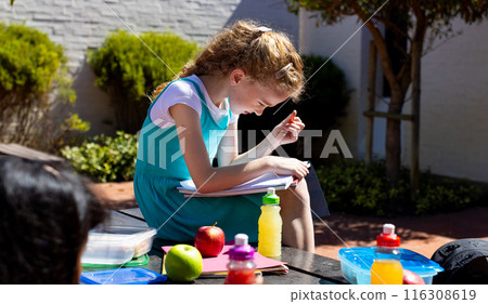 Caucasian girl with curly blonde hair is focused on reading outdoors in school Caucasian girl with curly blonde hair is focused on reading outdoors in school 116308619