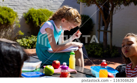 Caucasian girl focuses on her schoolwork outdoors, surrounded by peers Caucasian girl focuses on her schoolwork outdoors, surrounded by peers 116308620