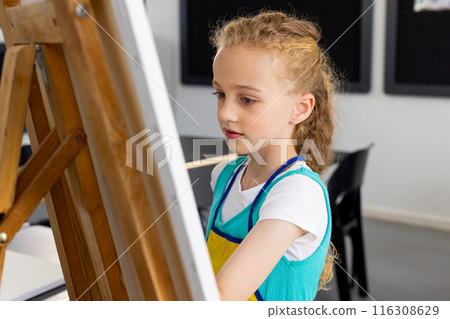 Caucasian girl with curly blonde hair is painting on an easel in school Caucasian girl with curly blonde hair is painting on an easel in school 116308629