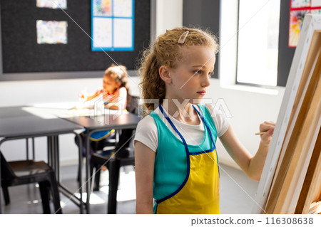 Caucasian girl with blonde hair paints on an easel, wearing a blue apron in school Caucasian girl with blonde hair paints on an easel, wearing a blue apron in school 116308638
