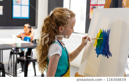 Caucasian girl paints with blue and yellow on an easel in school, biracial girl in the background 116308639