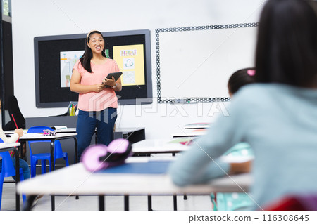In school, young biracial female teacher holding tablet stands smiling in classroom 116308645