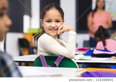 Young biracial girl with brown hair is sitting, smiling at her desk in classroom 116308691