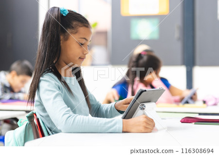 In school, young biracial girl holding tablet sits at a desk in classroom 116308694