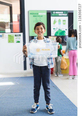 In school, biracial boy holding plastic recycling bin stands smiling in classroom 116308727