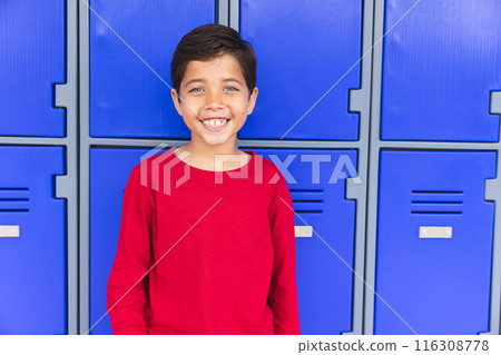 In school, young biracial male student standing in front of blue lockers outdoors, smiling 116308778