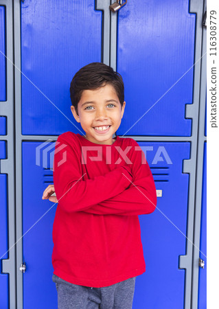 In school, young biracial male student leaning against blue lockers outdoors, arms crossed 116308779