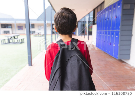 In school, biracial young boy wearing a red shirt and backpack walking outdoors 116308780
