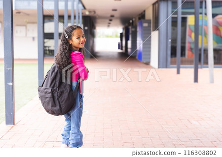In school, a young biracial girl wearing a backpack standing in a hallway outdoors with copy space In school, a young biracial girl wearing a backpack standing in a hallway outdoors with copy space 116308802