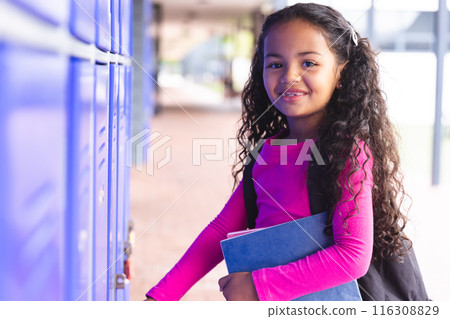 In school, young biracial female student holding books, standing by lockers outdoors In school, young biracial female student holding books, standing by lockers outdoors 116308829