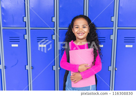 In school, young biracial girl holding a pink folder stands smiling with copy space beside lockers In school, young biracial girl holding a pink folder stands smiling with copy space beside lockers 116308830