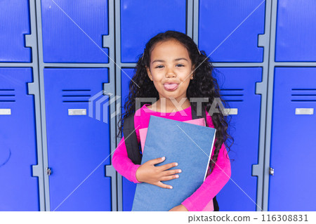 In school, young biracial girl holding a blue folder sticks out her tongue In school, young biracial girl holding a blue folder sticks out her tongue 116308831