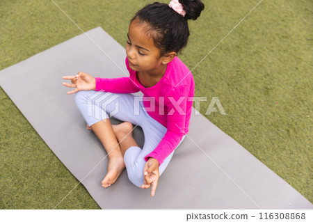 A young biracial girl wearing pink is sitting on a yoga mat outdoors 116308868