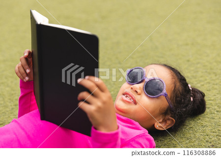 In school, young biracial girl wearing sunglasses lying on grass outdoors reading 116308886