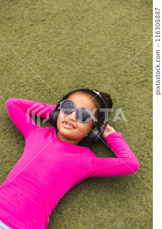 In school, young biracial girl is lying on grass outdoors, wearing headphones In school, young biracial girl is lying on grass outdoors, wearing headphones 116308887