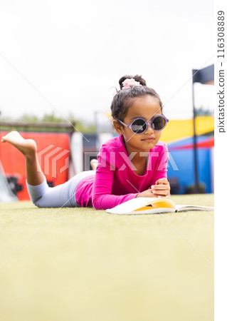 In school, young biracial girl wearing sunglasses is reading a book on grass outdoors 116308889