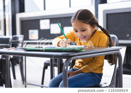 In school, young biracial girl with brown hair wearing a yellow top is writing in the classroom In school, young biracial girl with brown hair wearing a yellow top is writing in the classroom 116308924