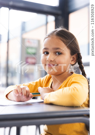 In school, young biracial girl holding a pencil, sitting at a desk in a classroom 116308929