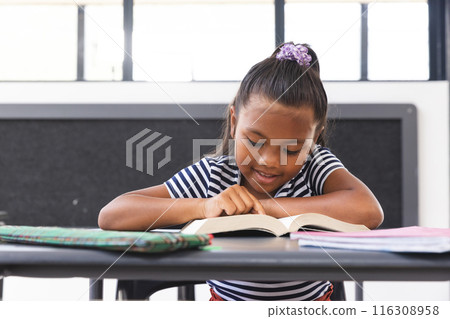 In school, young biracial girl is reading a book at her desk in the classroom In school, young biracial girl is reading a book at her desk in the classroom 116308958