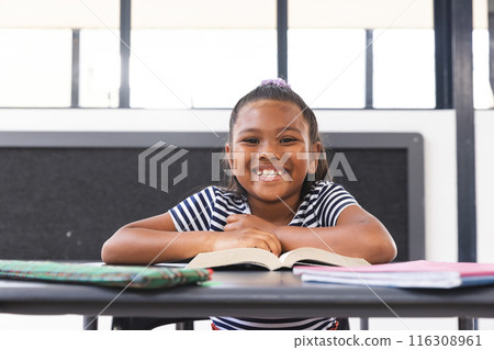 In school, young biracial girl is sitting at a desk in a classroom, reading a book 116308961