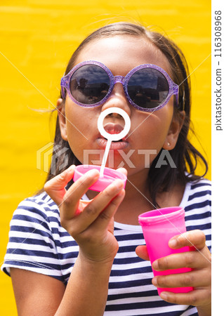 In school, young biracial girl wearing sunglasses is blowing bubbles outdoors 116308968