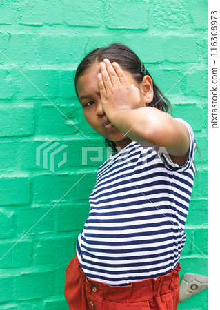 In school, young biracial girl is covering one eye with her hand outdoors 116308973