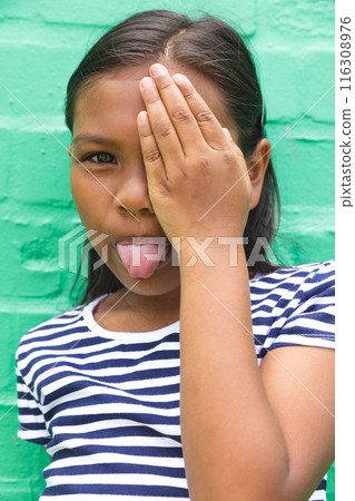 In school, outdoors, biracial young girl covering one eye, sticking out tongue In school, outdoors, biracial young girl covering one eye, sticking out tongue 116308976